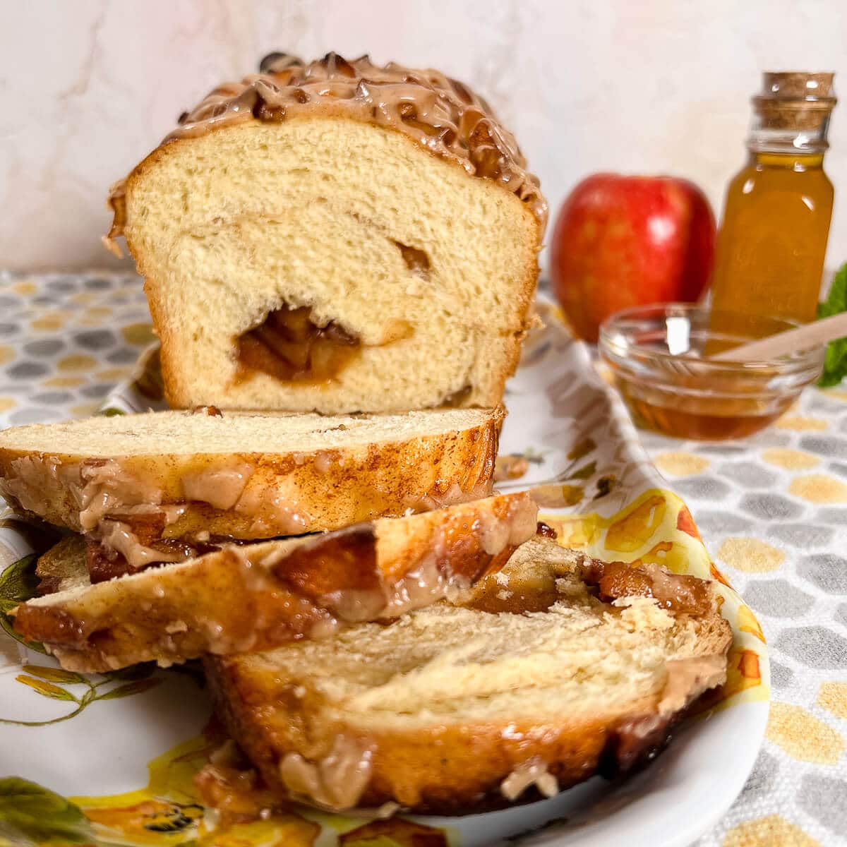 Sliced apple honey babka on a plate with an apple and honey in a jar and bowl beside all on a honeycomb-decorated towel.