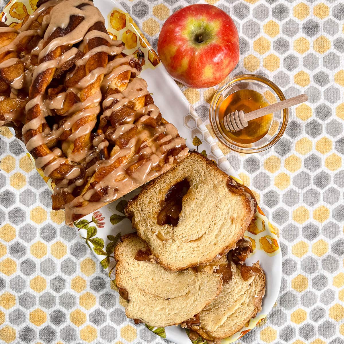 Sliced apple honey babka on a plate with an apple and honey in a bowl beside all on a honeycomb-decorated towel from overhead.