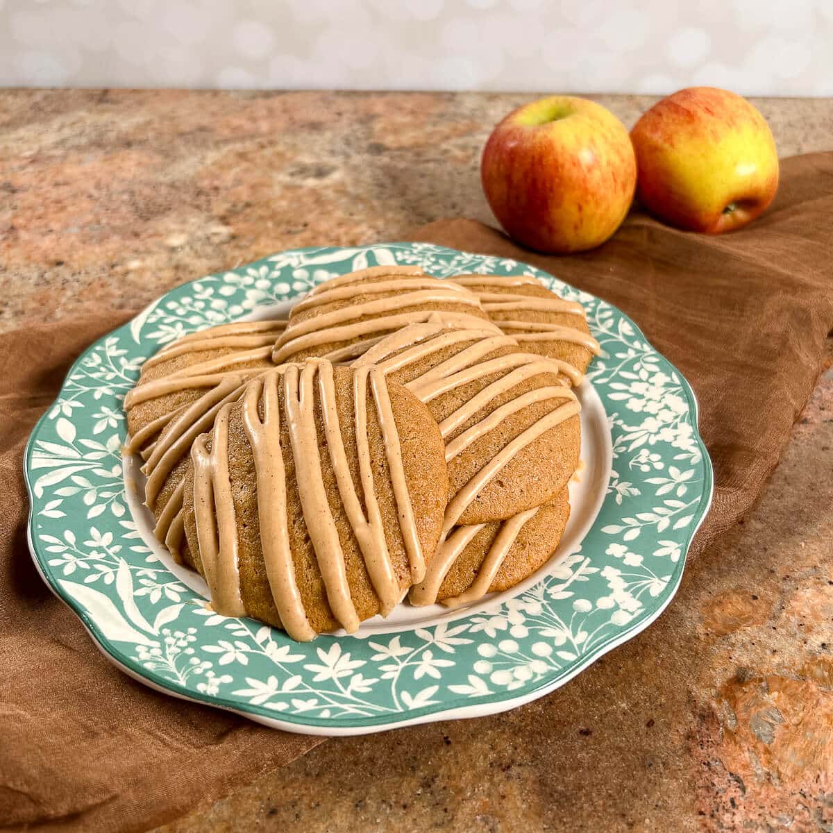 Apple spice cookies stacked on a plate with a green flowed edge and two apples behind.