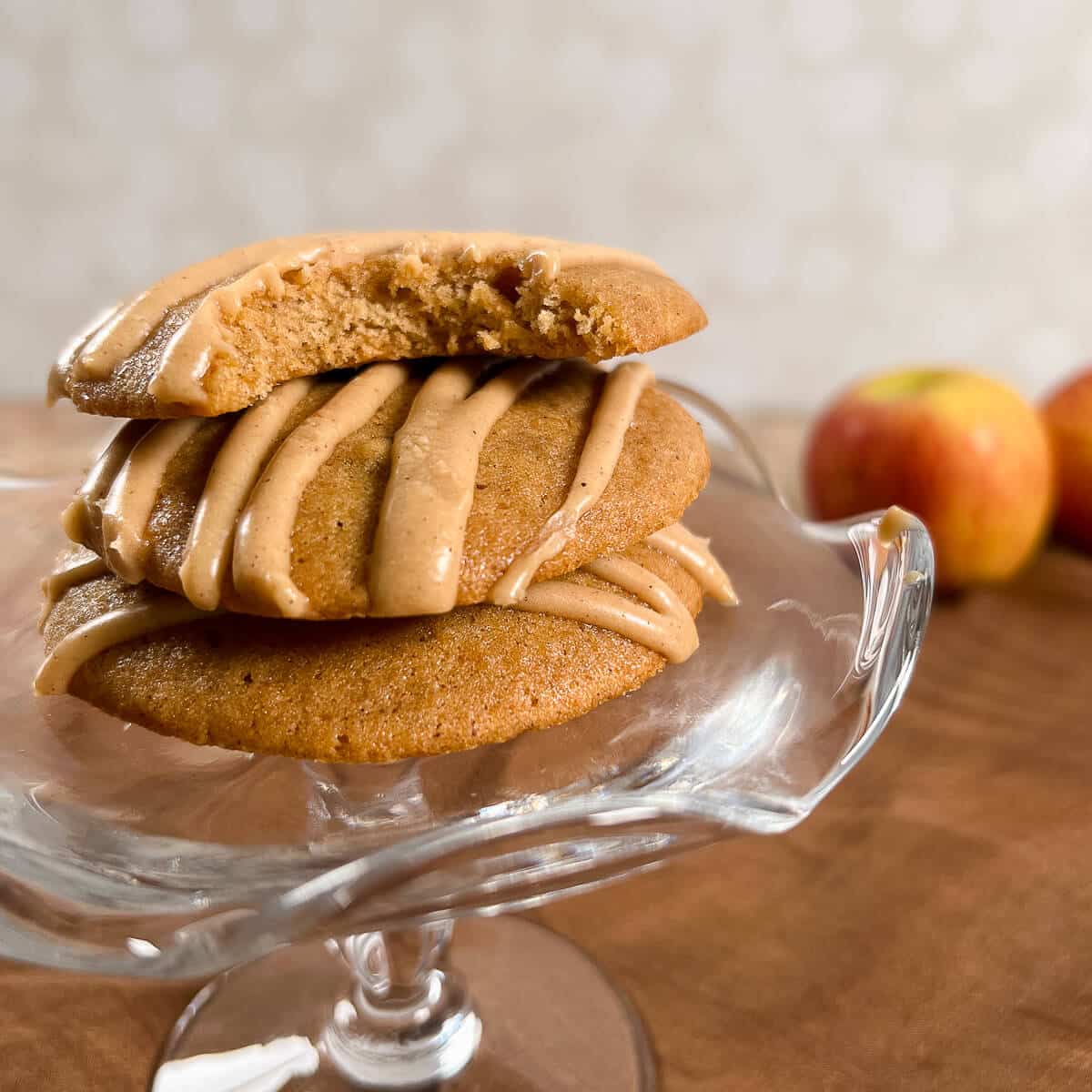 Apple spice cookies stacked on a glass stand with a bite out of the top one and apples behind.