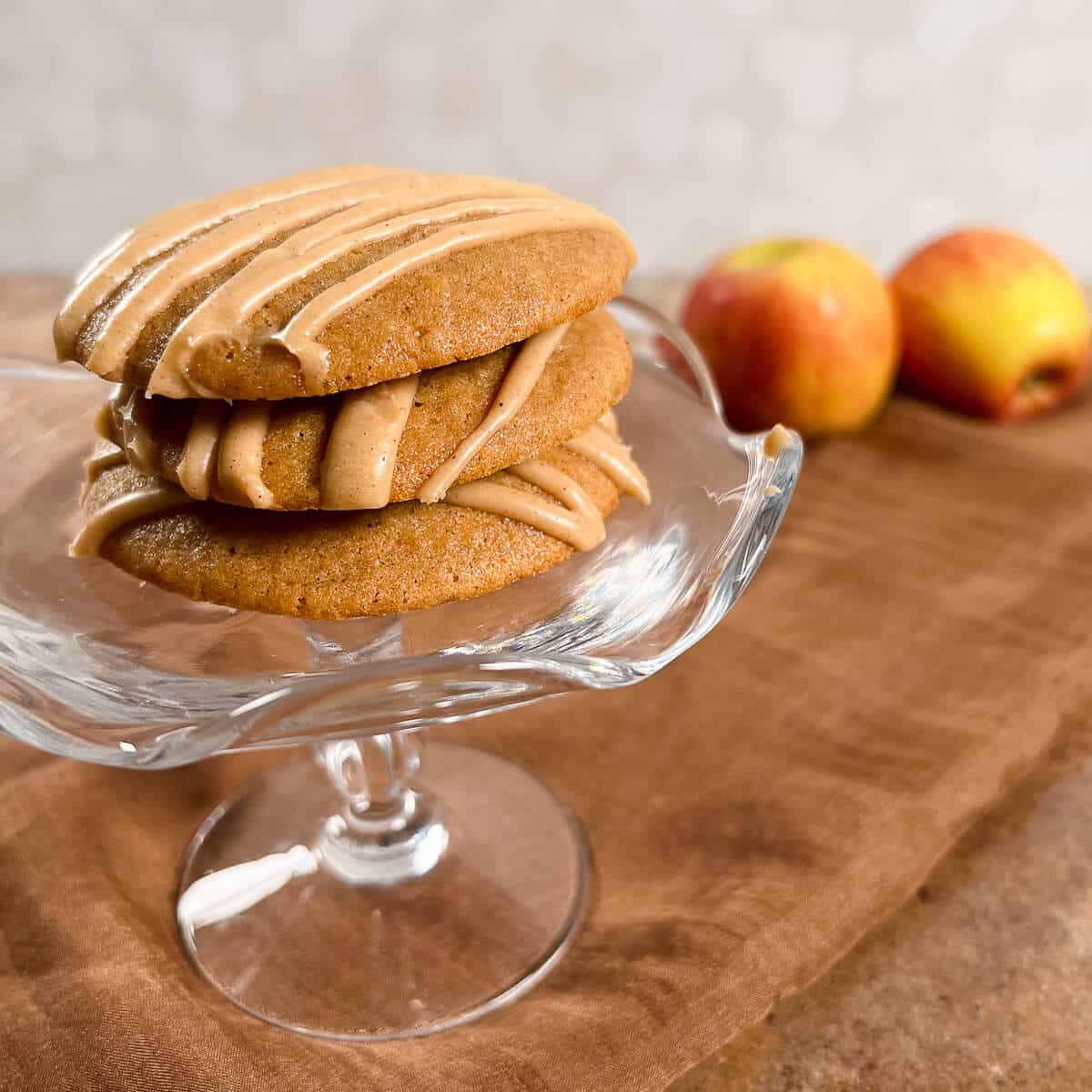 Apple spice cookies stacked on a glass stand with two apples behind.