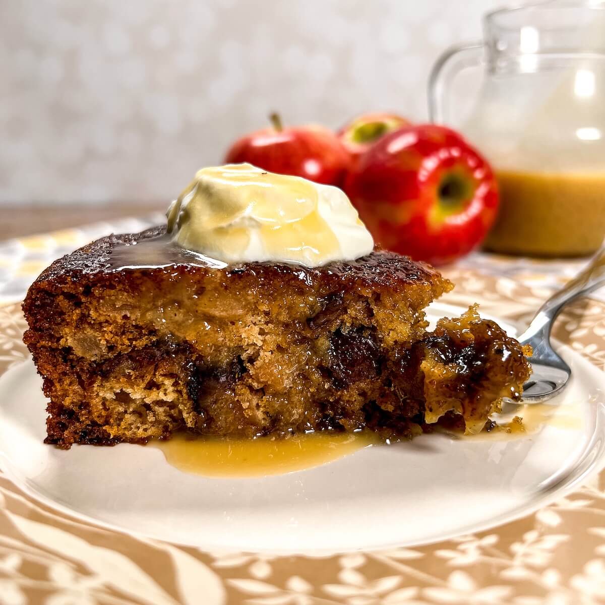 Slice of sticky toffee apple pudding with a bite on a fork on a plate with the rest of the cake on a cake stand with jug of honey toffee sauce and apples behind.