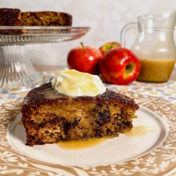 Slice of sticky toffee apple pudding on a plate with the rest of the cake on a cake stand with jug of honey toffee sauce and apples behind.