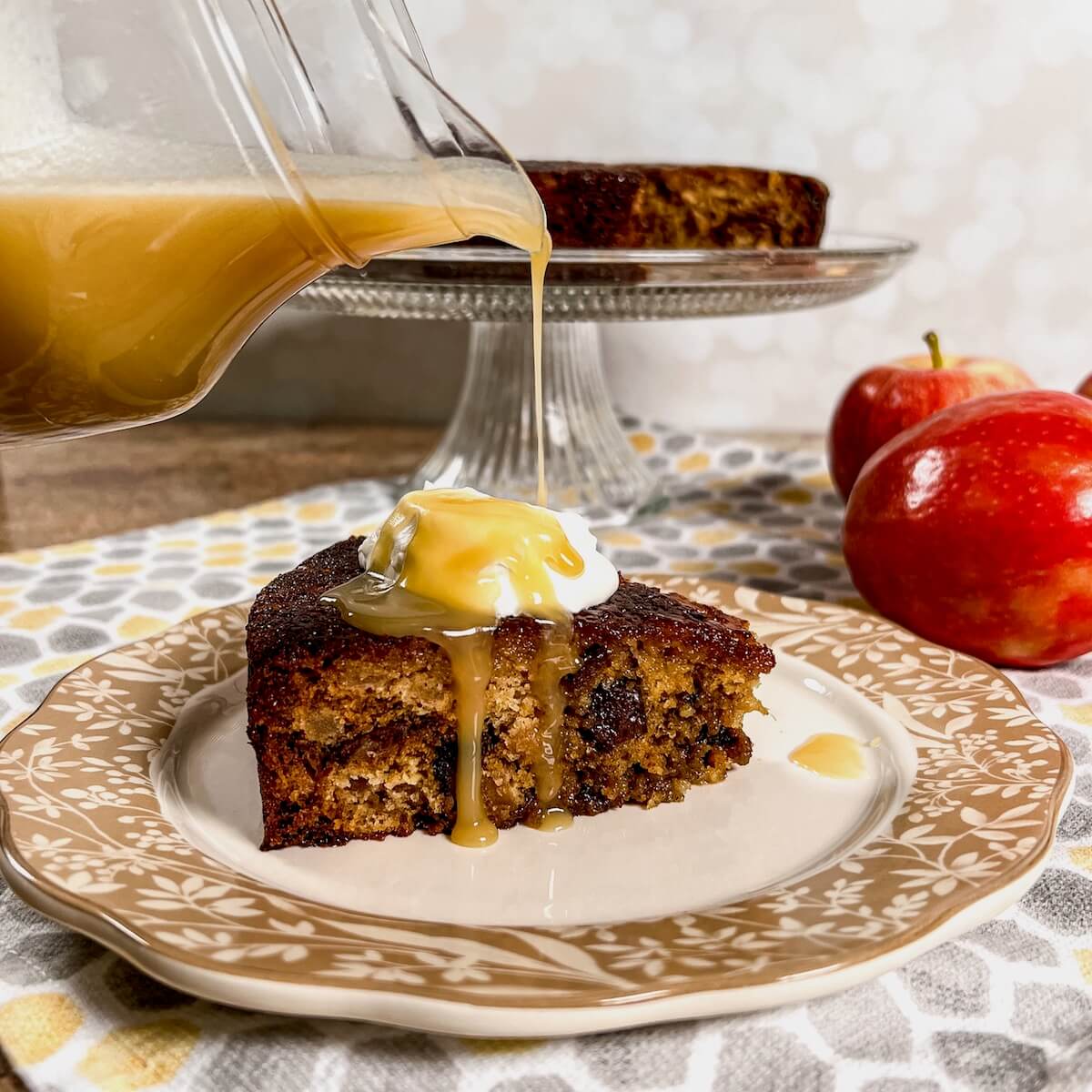 Pouring honey toffee sauce onto a slice of sticky toffee apple pudding on a plate with the rest of the cake on a cake stand and apples behind.