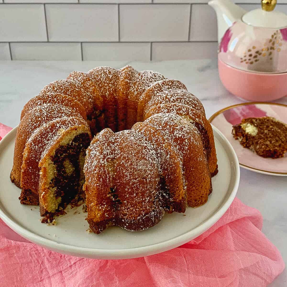 Marble Bundt Cake on cake stand sliced with piece on plate & teapot in background.