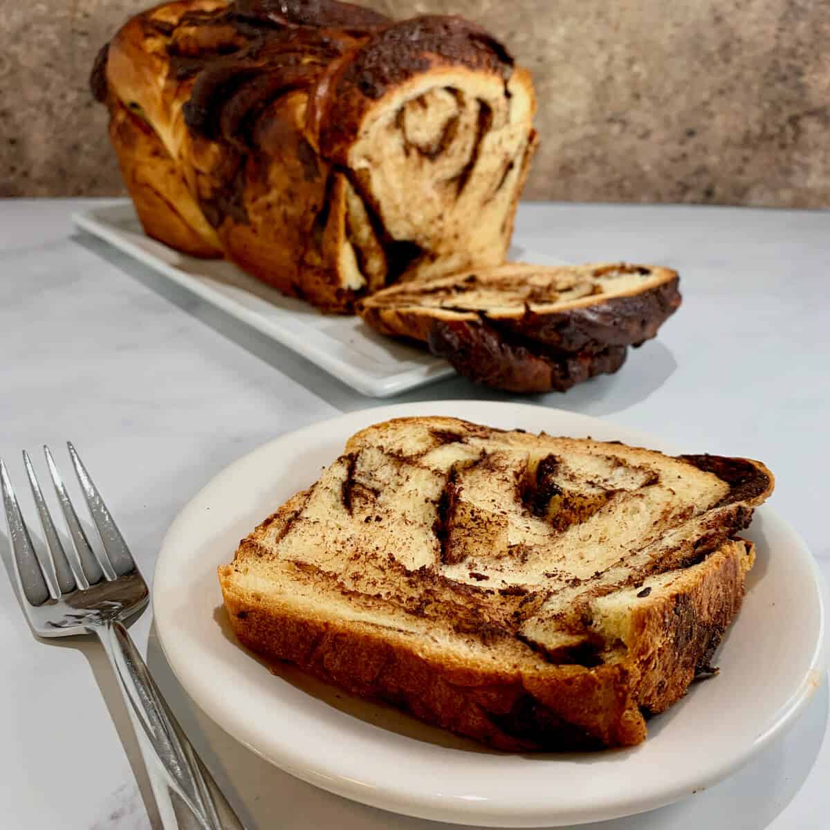 Slice of Chocolate Fudge Babka on a white plate with a fork & the loaf behind.