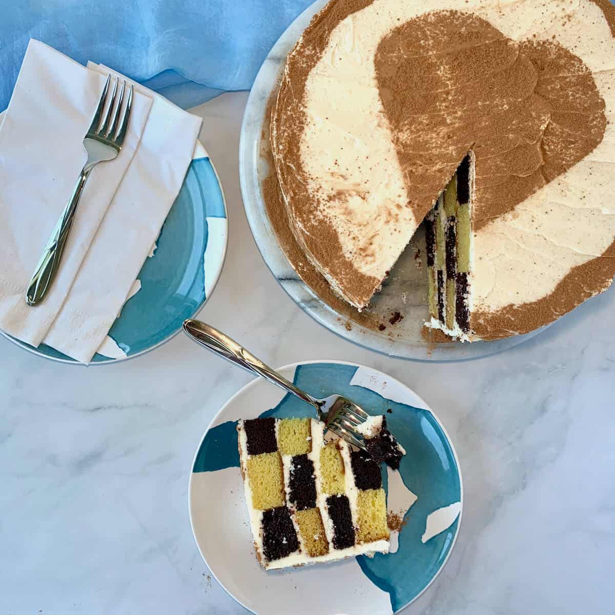 Checkerboard cake slice on a blue & white plate with cake on plate next to a fork.