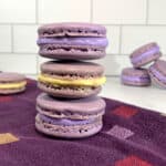 French Macarons with purple buttercream & lemon curd stacked on a purple towel with more in background.