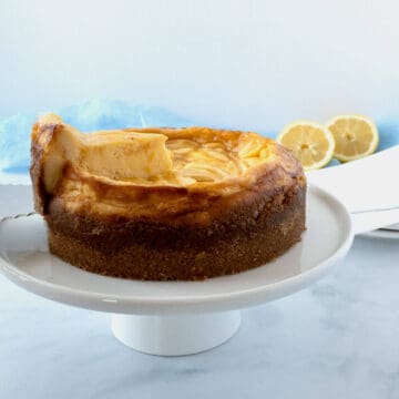 Lifting slice of Lemon Curd Marbled Cheesecake slice from cheesecake on white cake stand & lemons in background.