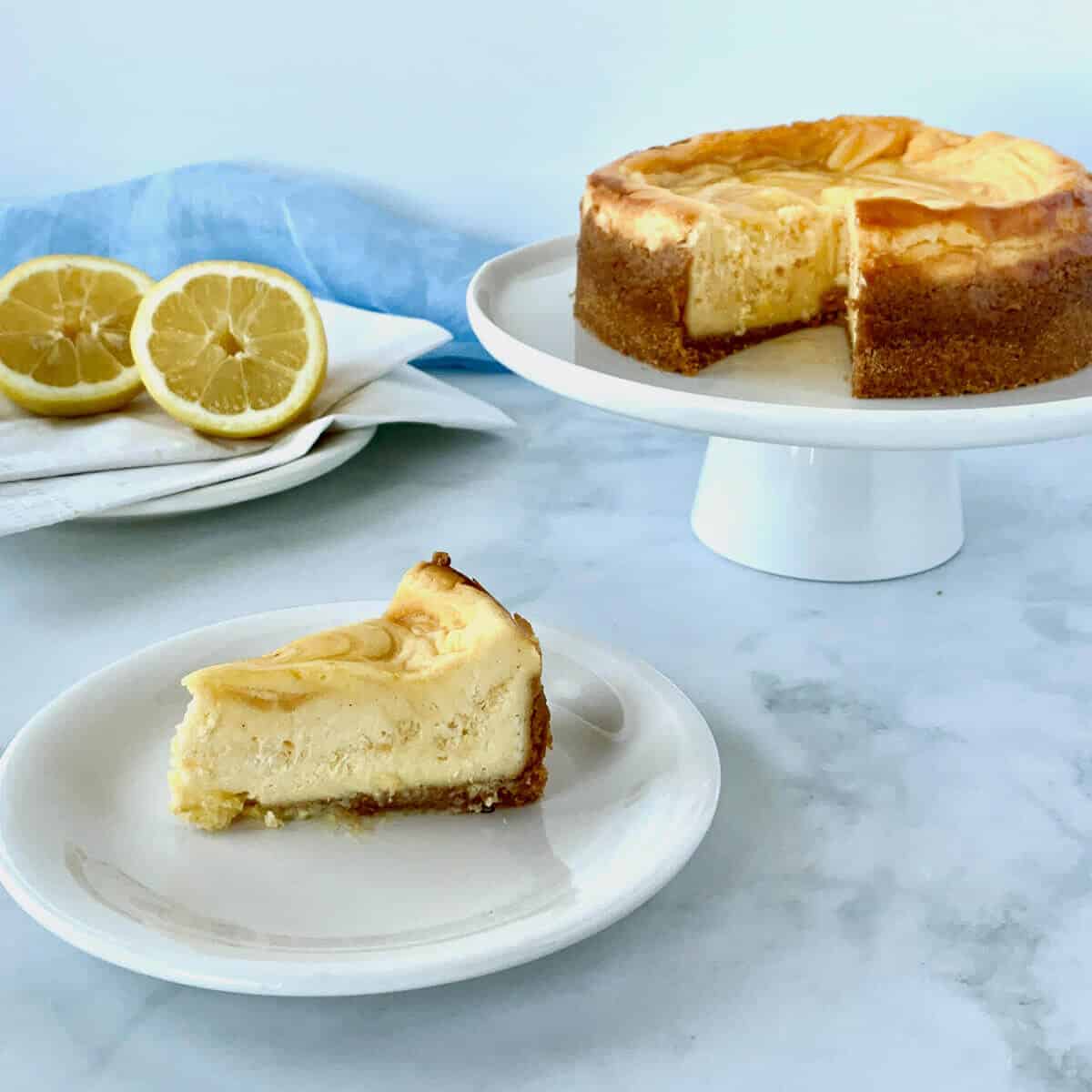 Lemon Curd Marbled Cheesecake slice on white plate with cheesecake on white cake stand & forks & lemons in background.