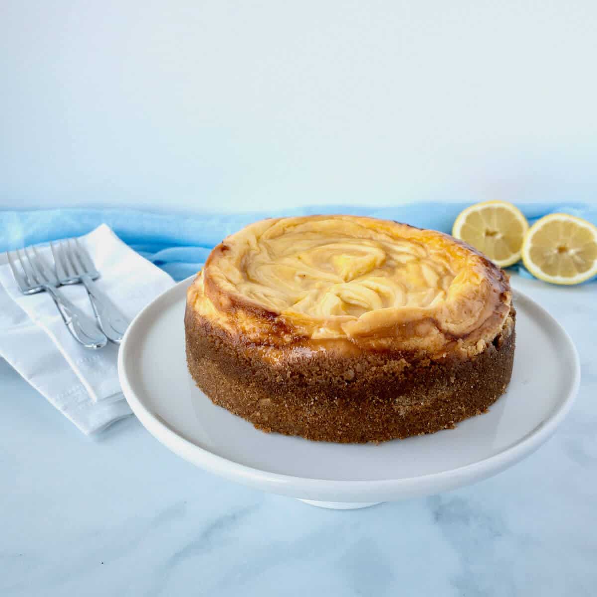 Lemon Curd Marbled Cheesecake on white cake stand with forks & lemons in background.