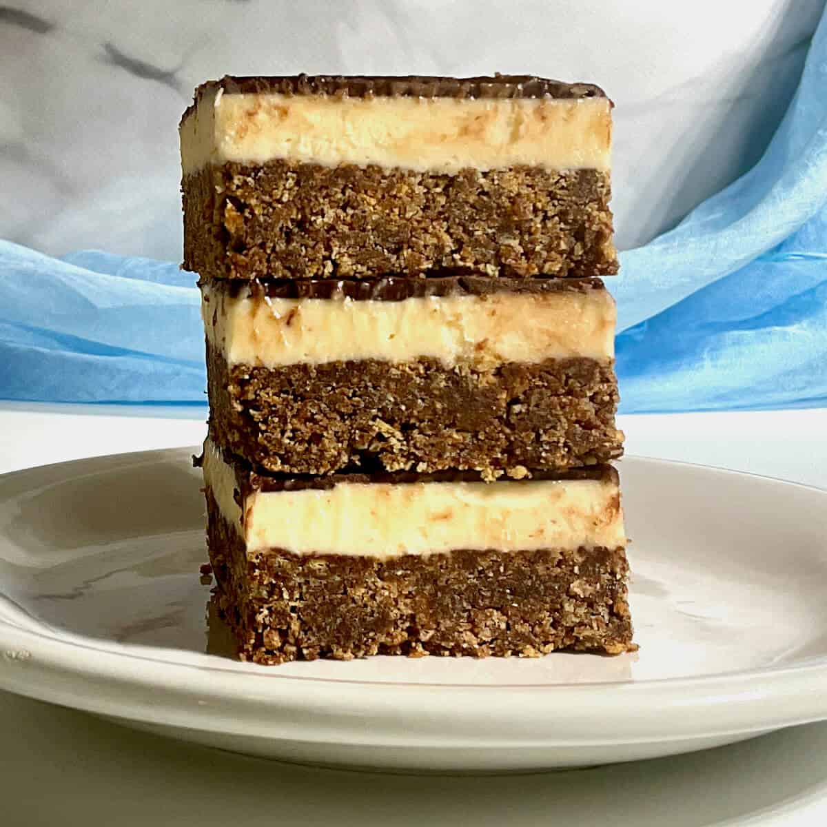 Nanaimo bars stacked on a white plate in front of a blue draped scarf.