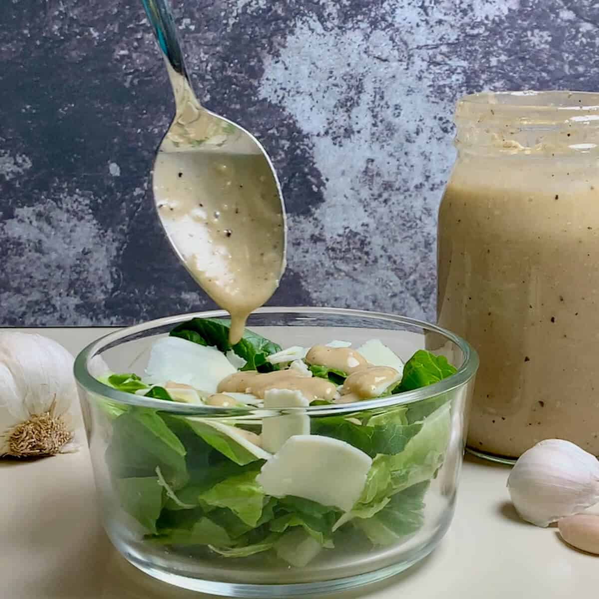 Drizzling Caesar dressing from a spoon onto a green salad with jar of dressing behind.