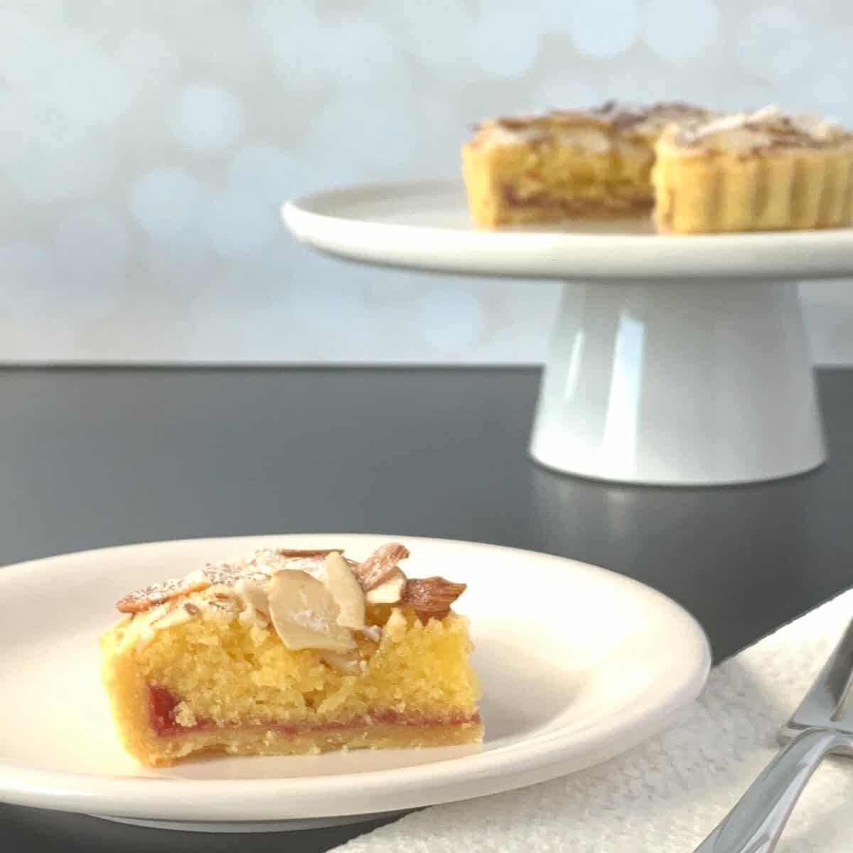 Slice of Bakewell tart on a white plate in front of a sliced tart on a white cakestand.