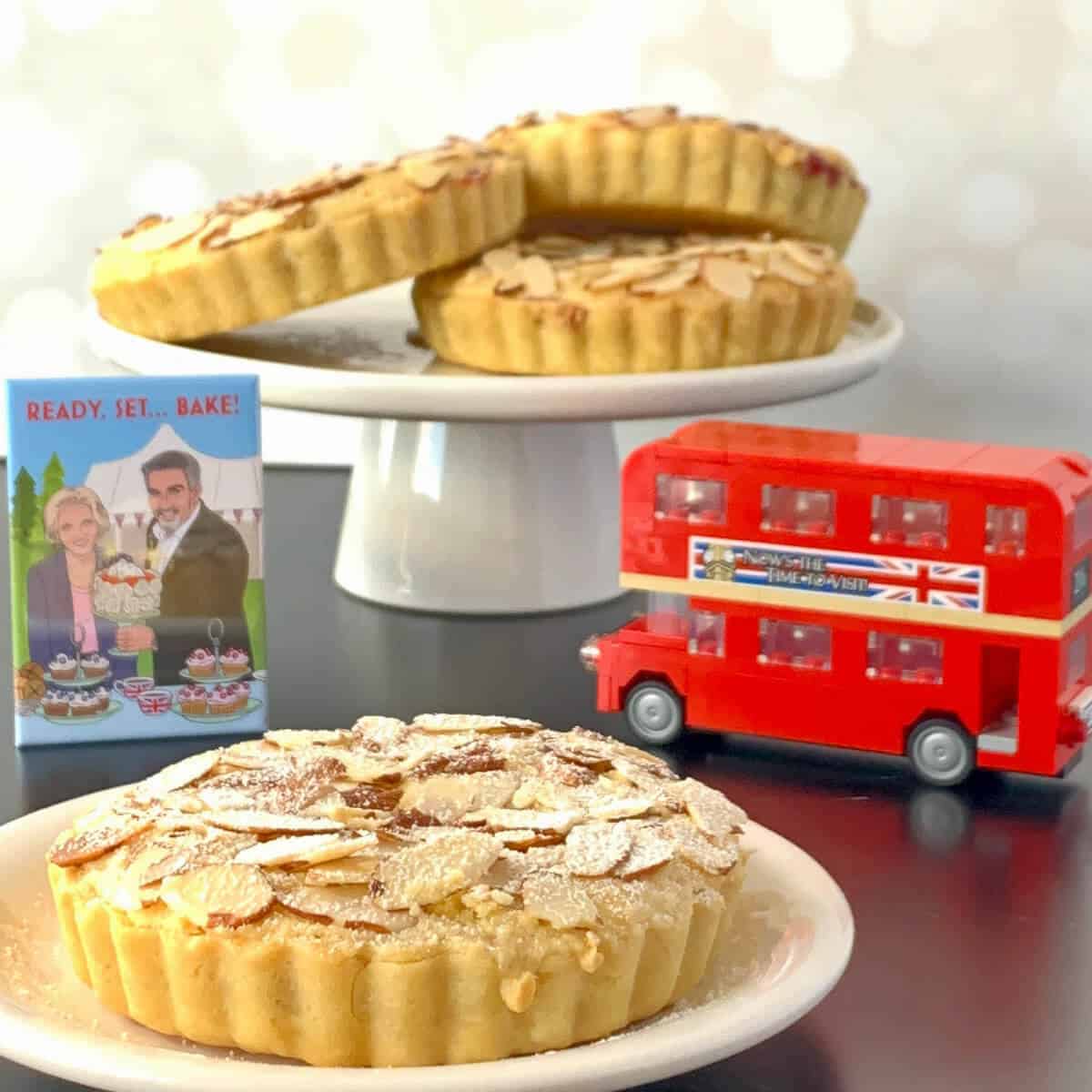 Mini Bakewell Tart on a white plate in front of 3 tarts on a white cake stand, a toy double-decker bus, & a Great British Bakeoff magnet.