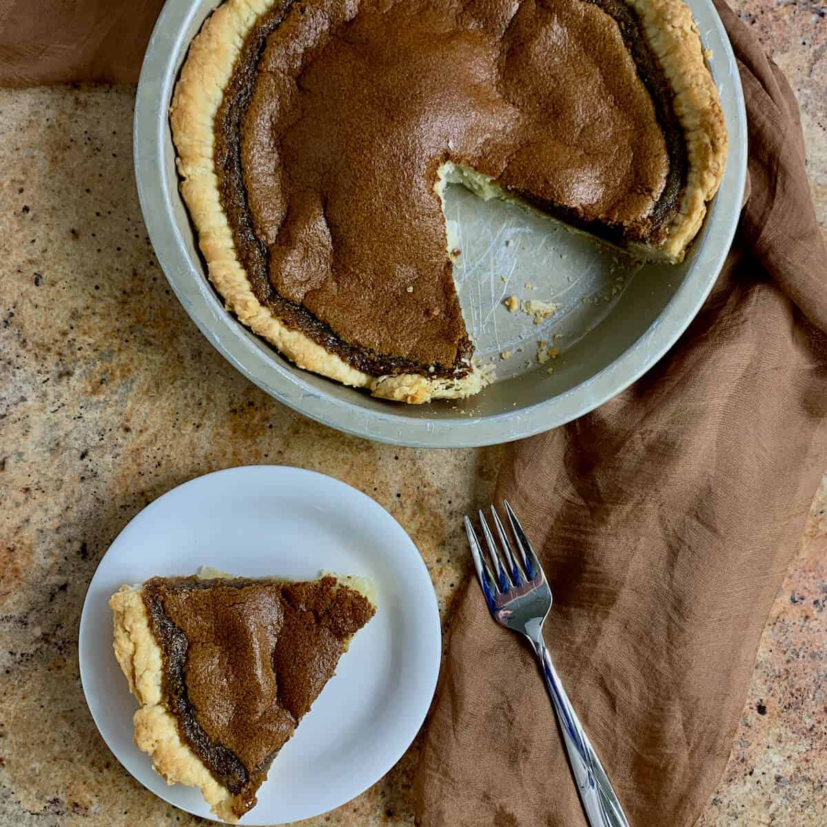 Slice of Brown Sugar Pie on a white plate and fork with the sliced pie in the pie pan on a brown scarf from overhead.