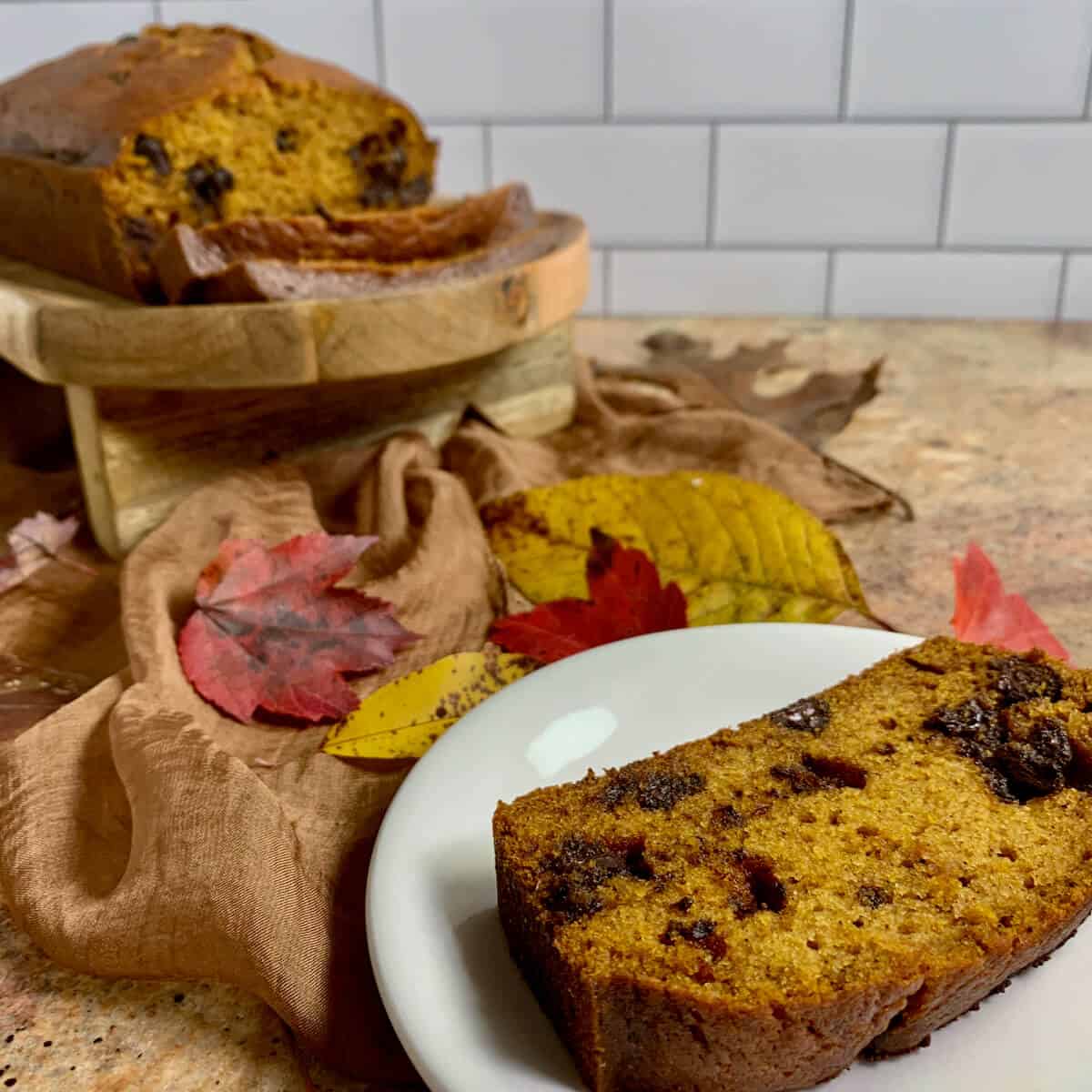Plated slice of Pumpkin Bread on a white plate with sliced loaf wood tray surrounded by Fall leaves on a brown scarf in background.