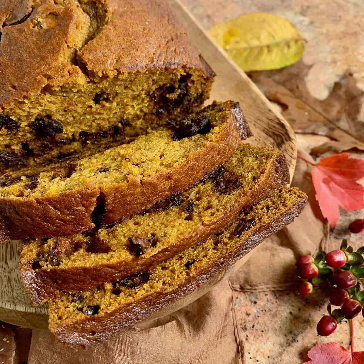 Closeup of sliced Pumpkin Bread on a wood tray surrounded by Fall leaves on a brown scarf.