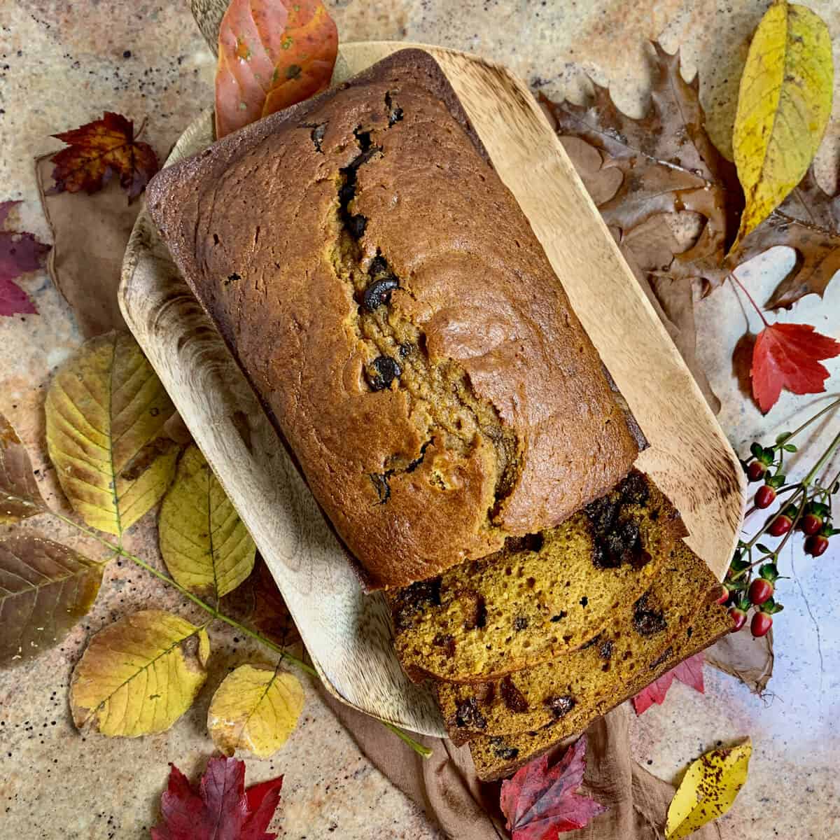 Sliced Pumpkin Bread on a wood tray surrounded by Fall leaves on a brown scarf from overhead.