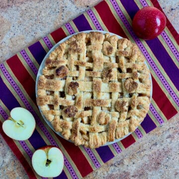 Lattice topped apple pie on a purple striped towel with cut apples from overhead.