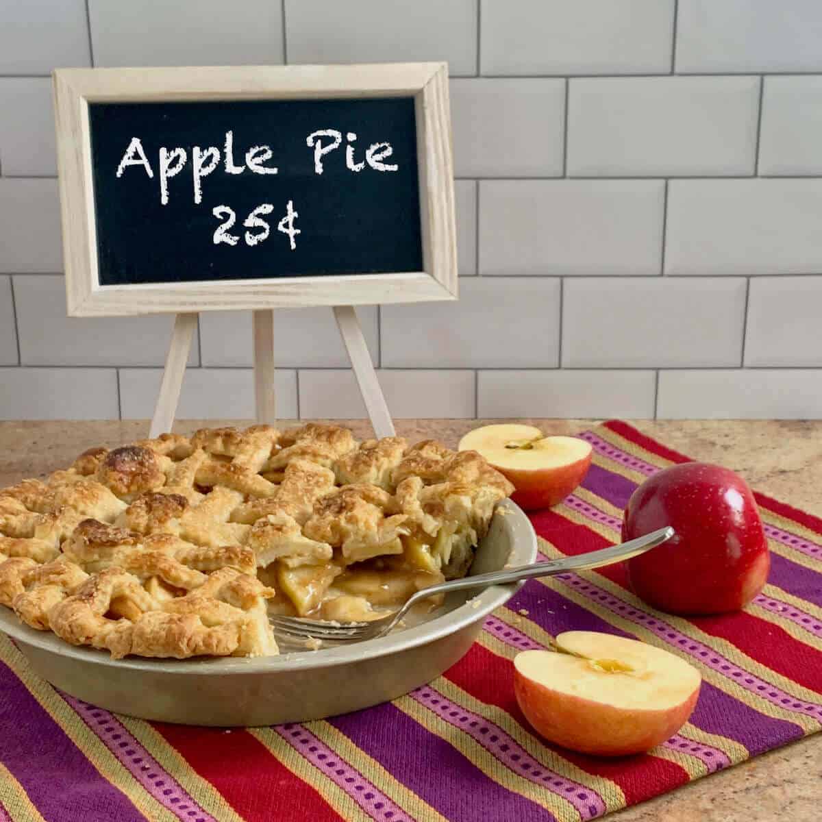 Sliced Apple Pie in pan with fork, apples, & chalkboard sign on a purple striped towel.