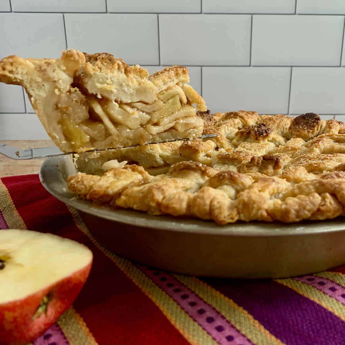 Apple Pie in pan with lifted slice & apples on a purple striped towel.