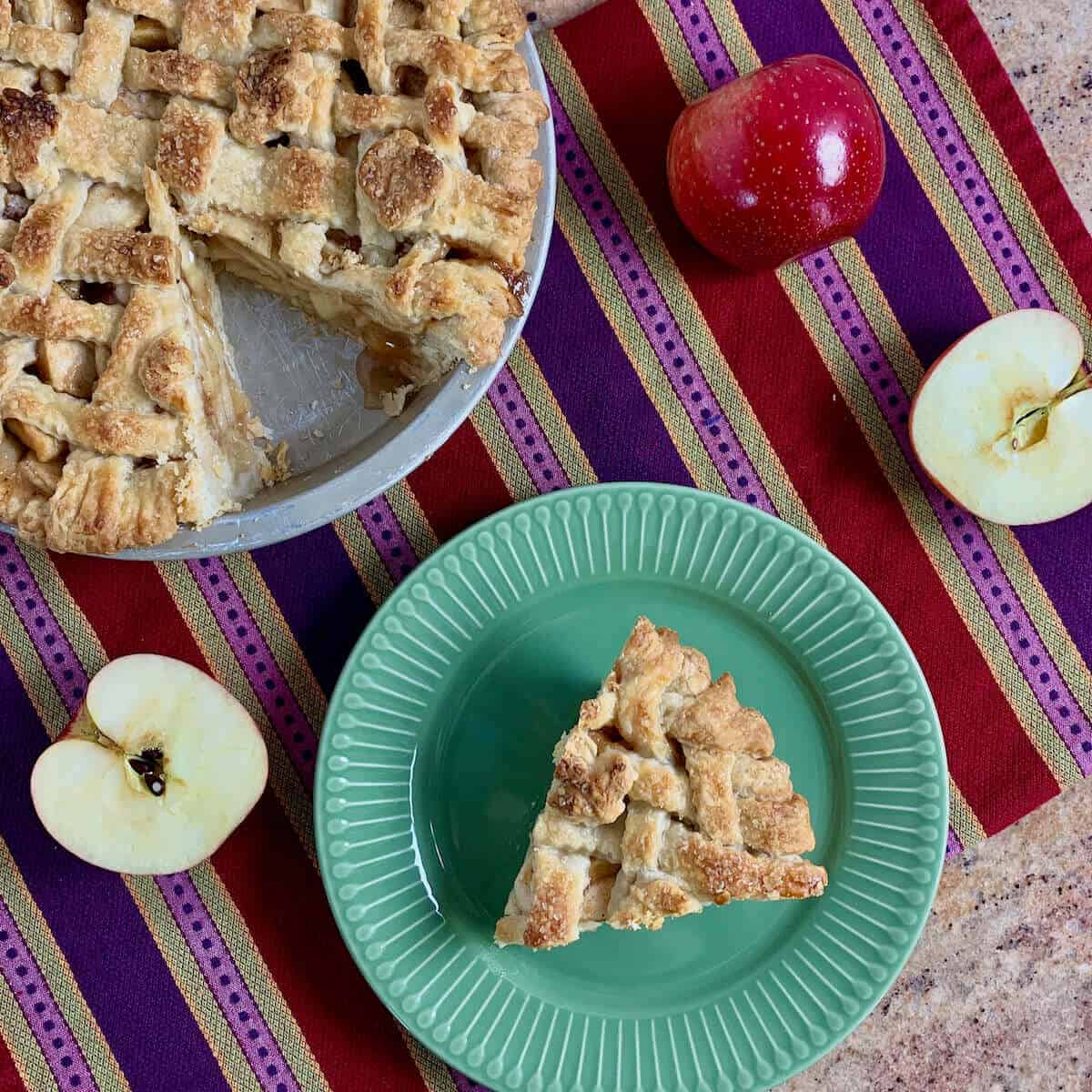 Slice of apple pie on a green plat with apples and whole sliced pie on a purple striped towel from overhead.