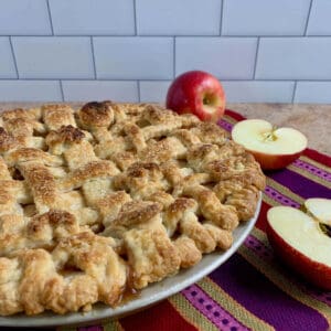 Lattice topped apple pie on a purple striped towel with cut apples.