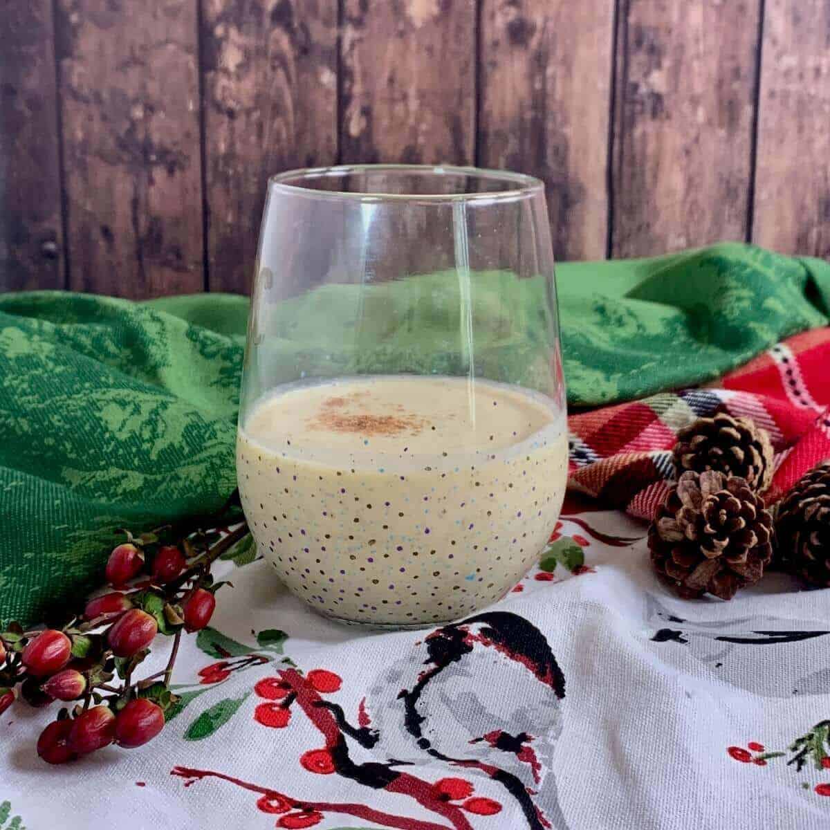 Glass of eggnog with festive greenery on a purple checked towel.