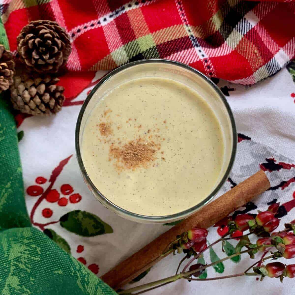 Glass of eggnog with cinnamon stick and pinecones on top of festive towels from overhead.