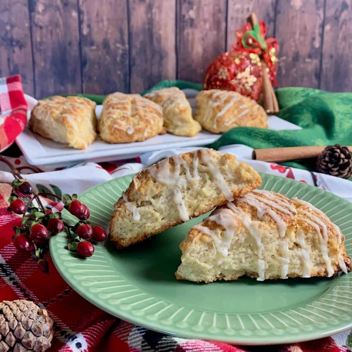 Two Eggnog Scones alongside red berries on a green plate with a white plate of 4 eggnog scones in the background.