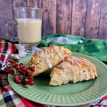Two Eggnog Scones alongside red berries on a green plate with a mug of eggnog in the background.