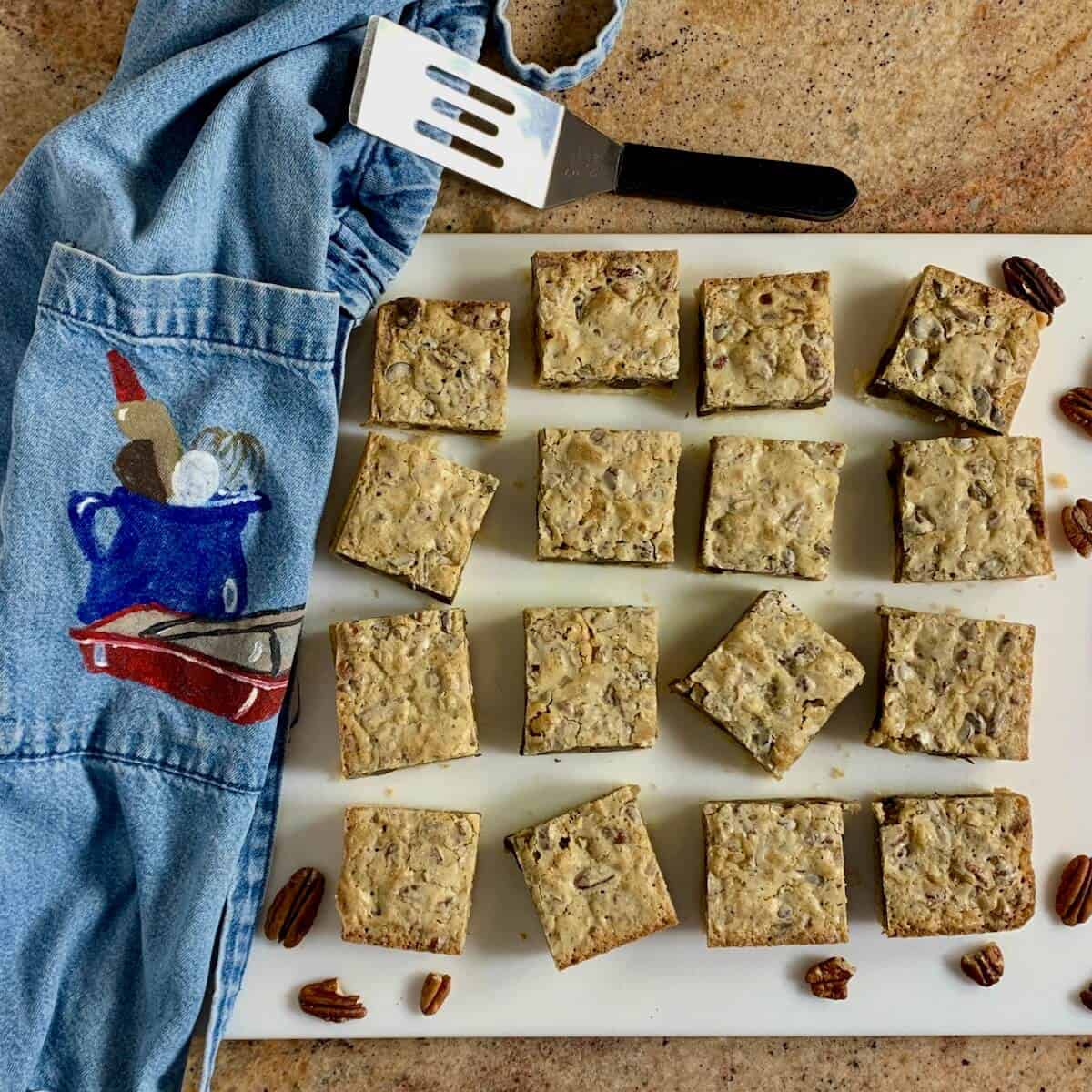 Pecan bars on a cutting board in a random pattern with a denim apron, pecans, and a small spatula from overhead.