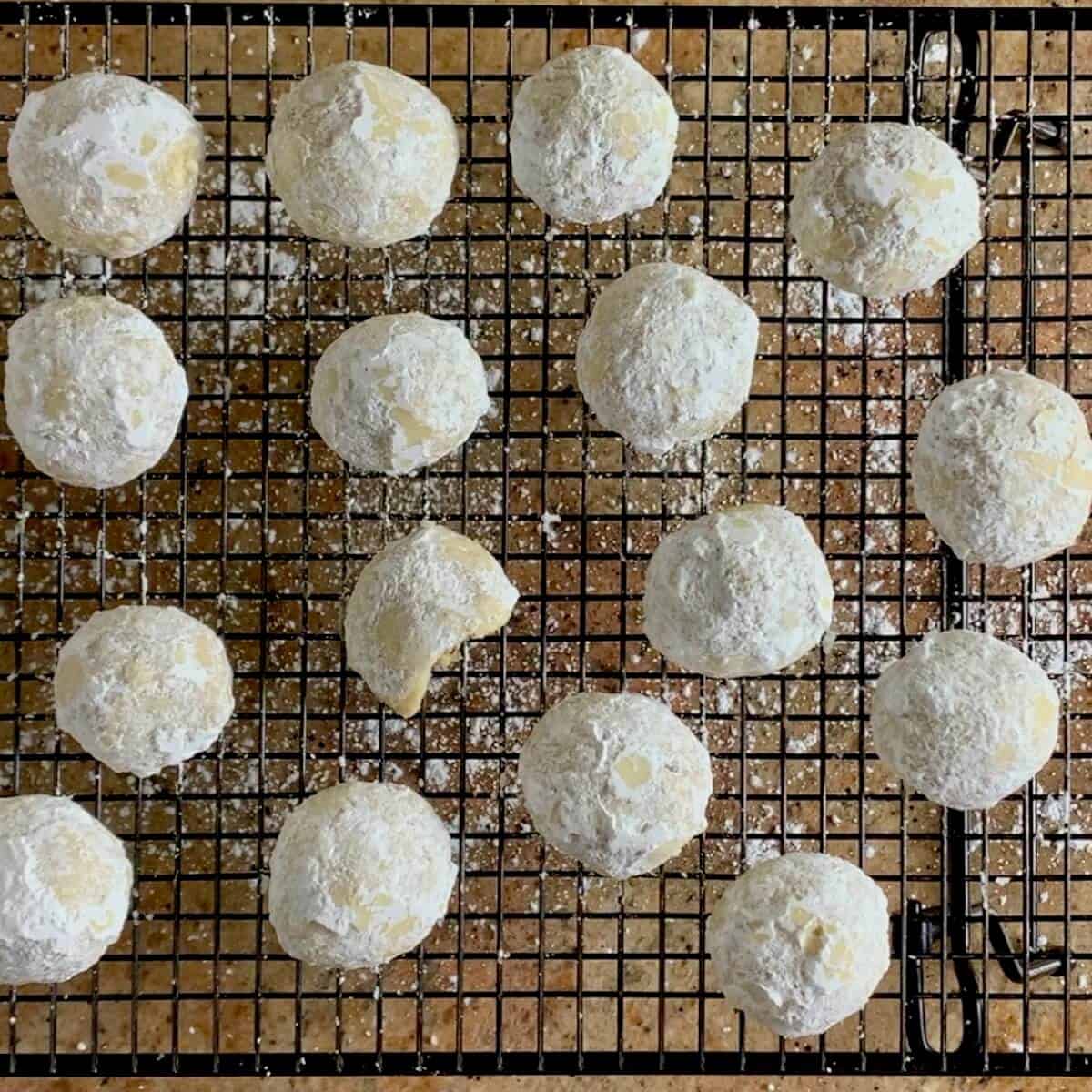 Finished batch of pecan snowball cookies on a cooling rack with one bitten.