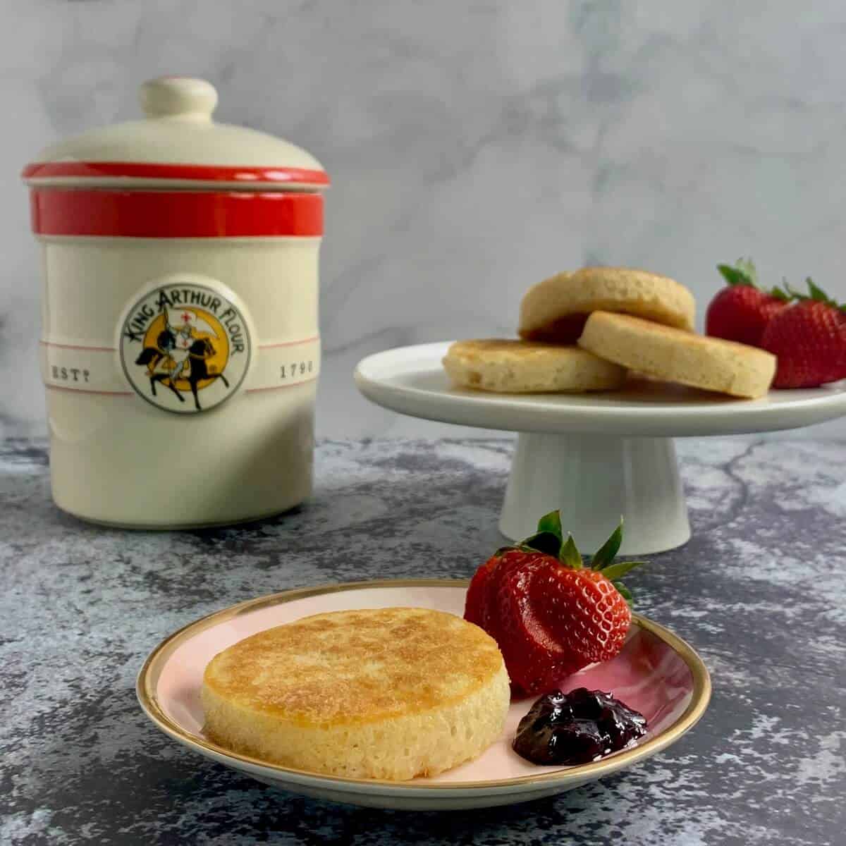 Crumpet on a plate with a sliced strawberry and jam in front of sourdough crock and a cakestand with 3 crumplets and some strawberries.