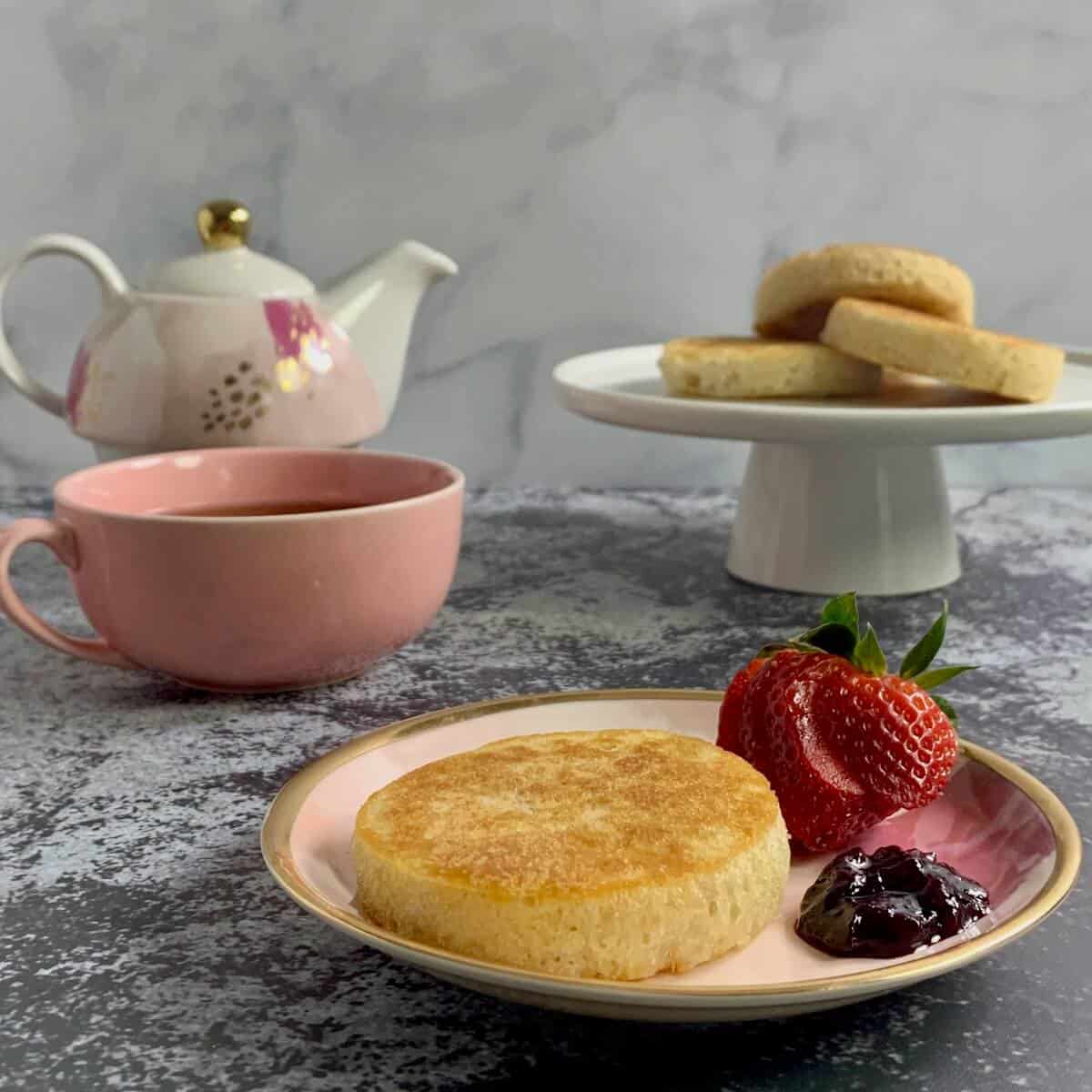 Crumpet on a plate with a sliced strawberry and jam in front of a cakestand with 3 crumplets and a teapot and pink teapot.