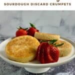 Three crumpets stacked on a white cakestand surrounded by strawberries Pinterest banner.