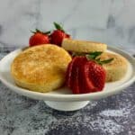 Three crumpets stacked on a white cake stand surrounded by strawberries.
