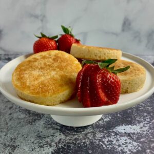Three crumpets stacked on a white cake stand surrounded by strawberries.