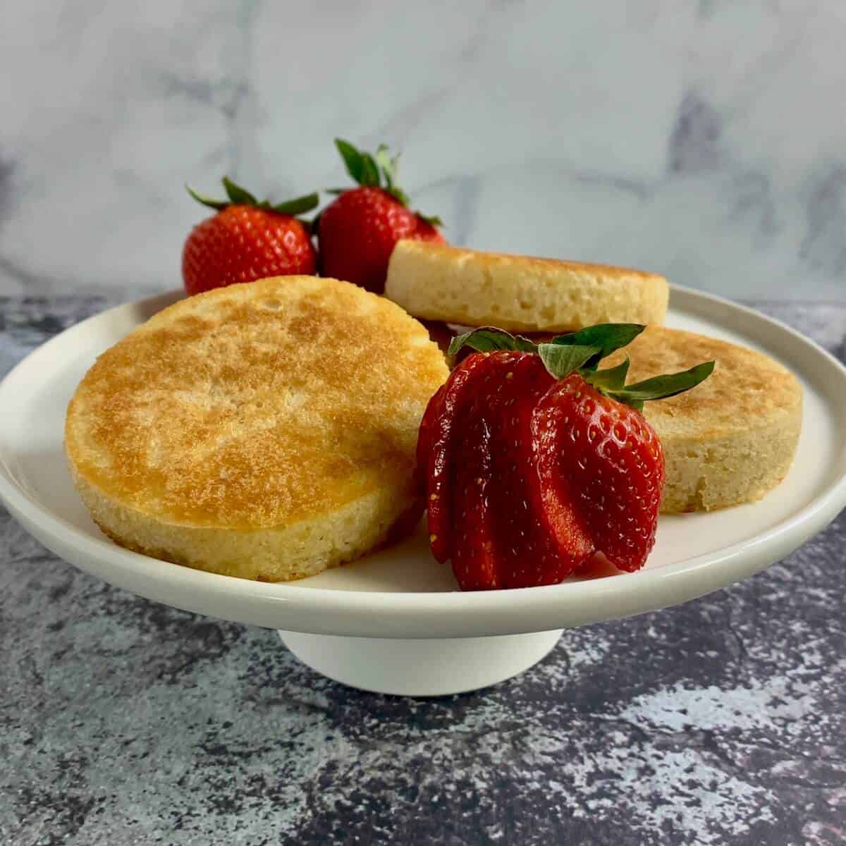 Three crumpets stacked on a white cakestand surrounded by strawberries.