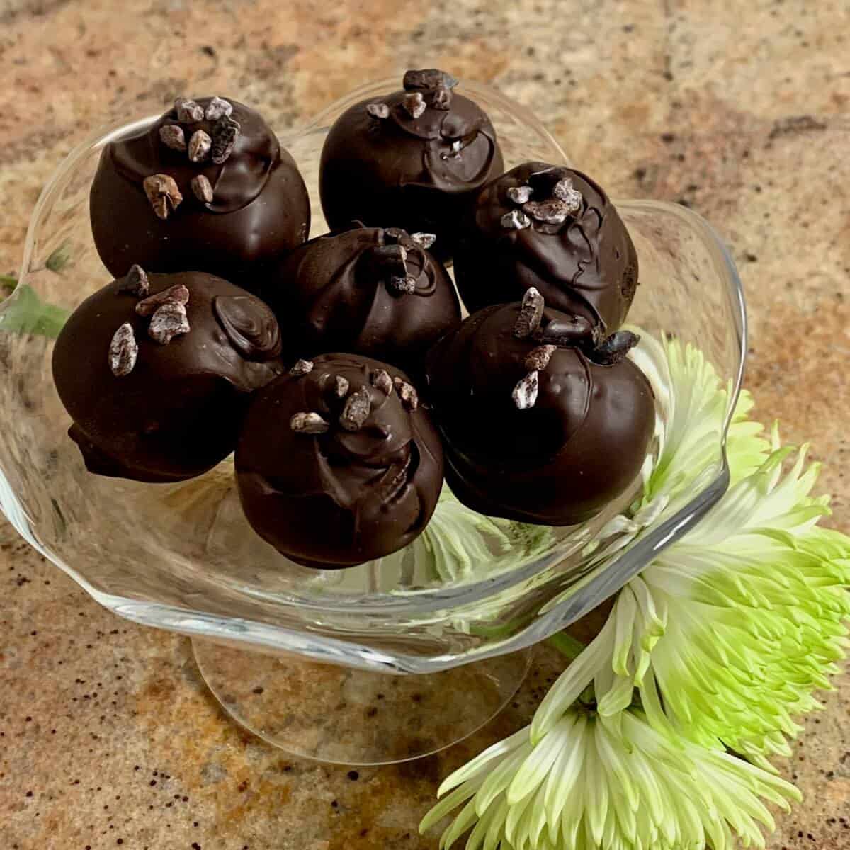 Chocolate cake truffles topped with cocoa nibs stacked on a glass plate stand with white flowers below from overhead.