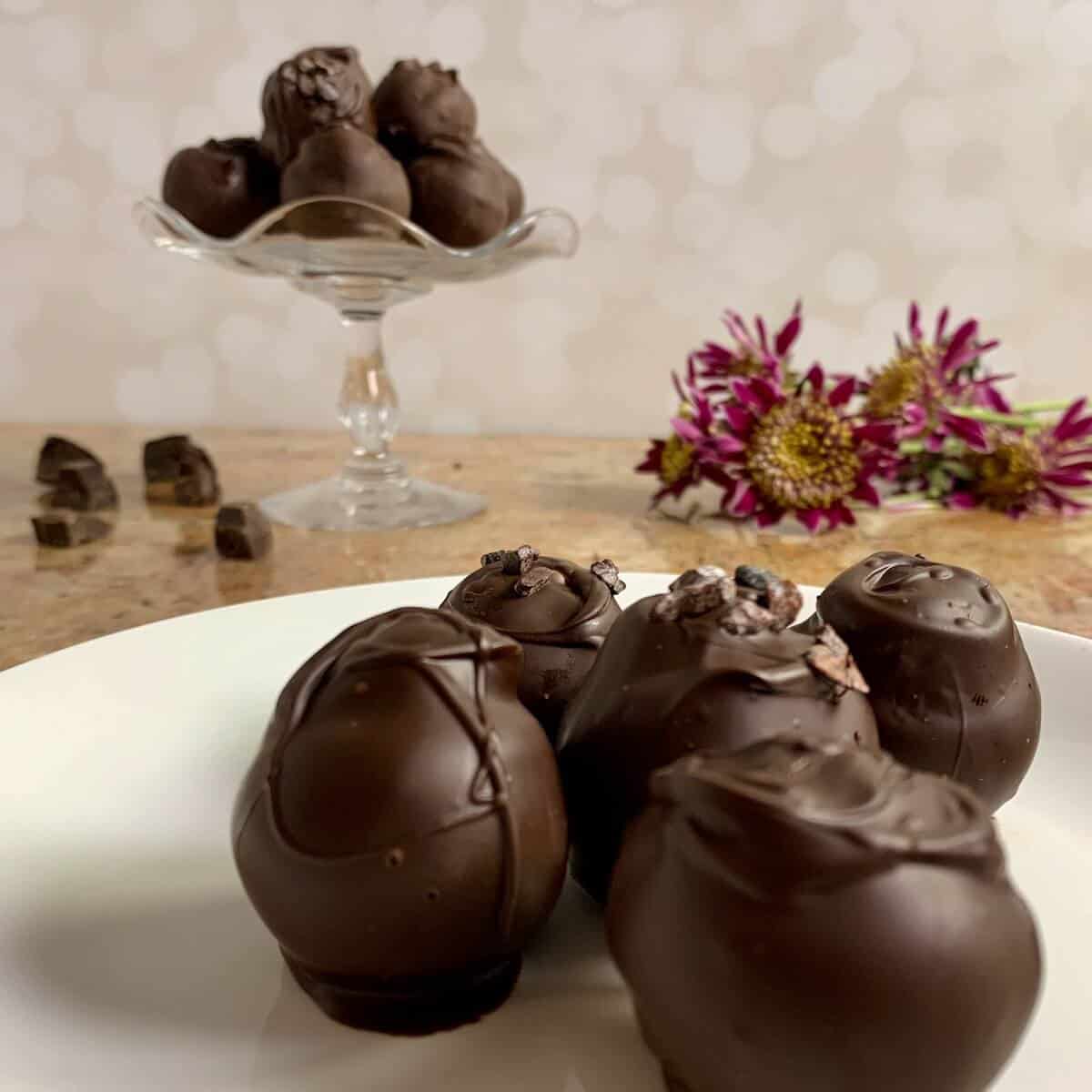 Chocolate cake truffles on a white plate in front of some stacked on a glass cake stand with pink flower.