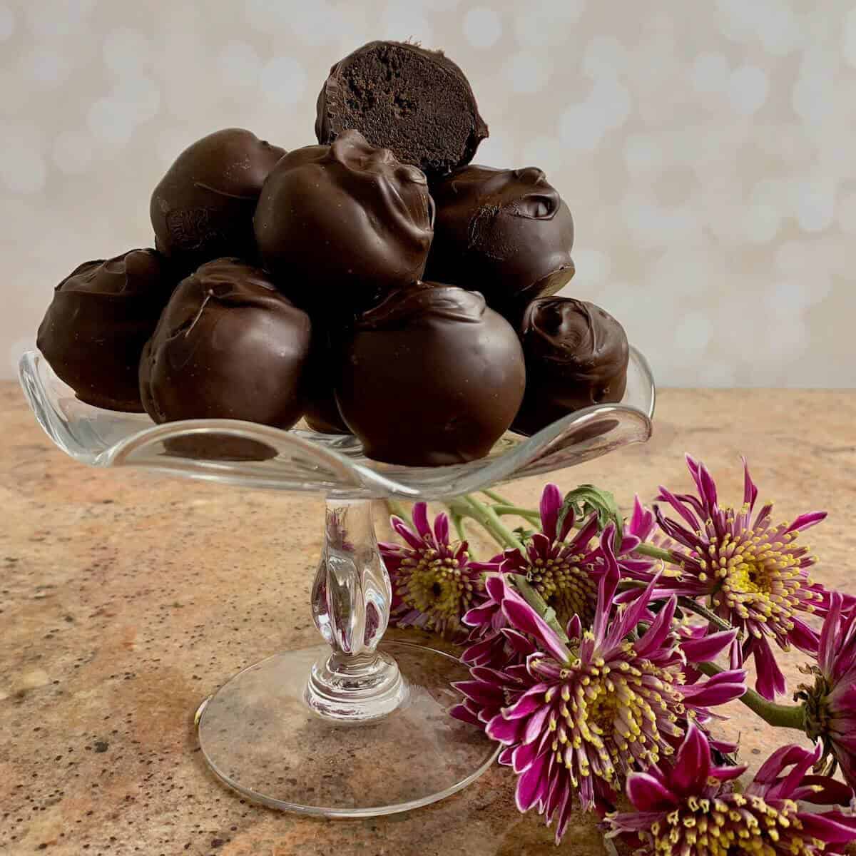 Chocolate cake truffles stacked on a glass plate stand with the top one cut and pink flowers below.