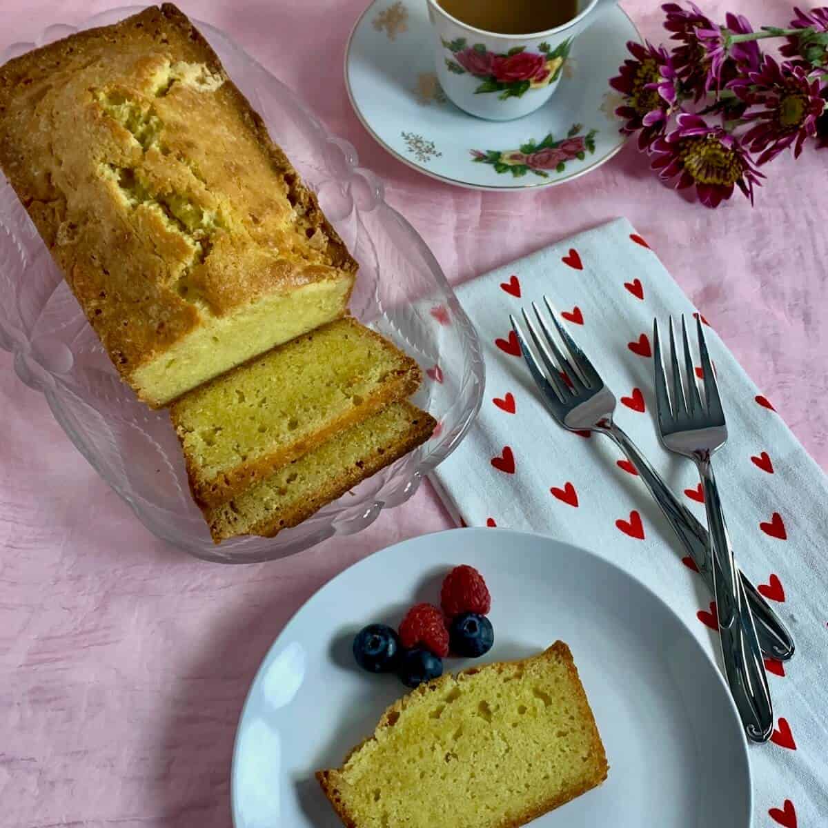 Pound cake slice on a white plate with sliced pound cake on a glass plate with a china coffee cup, forks, a heart spotted towel, and flowers.