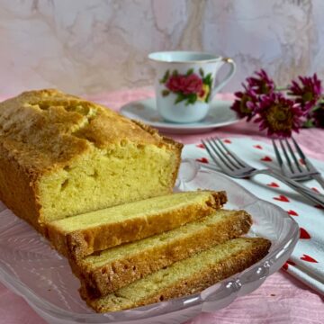 Sliced pound cake on a glass plate with a china coffee cup, forks, a heart spotted towel, and flowers.
