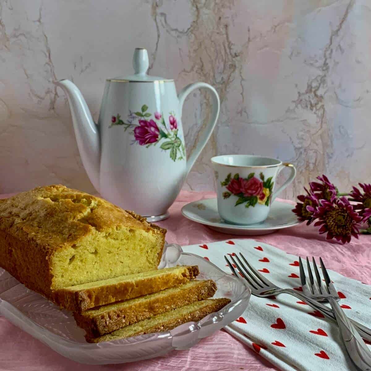 Sliced pound cake on a glass plate with a china pot and coffee cup, forks, a heart spotted towel, and flowers.