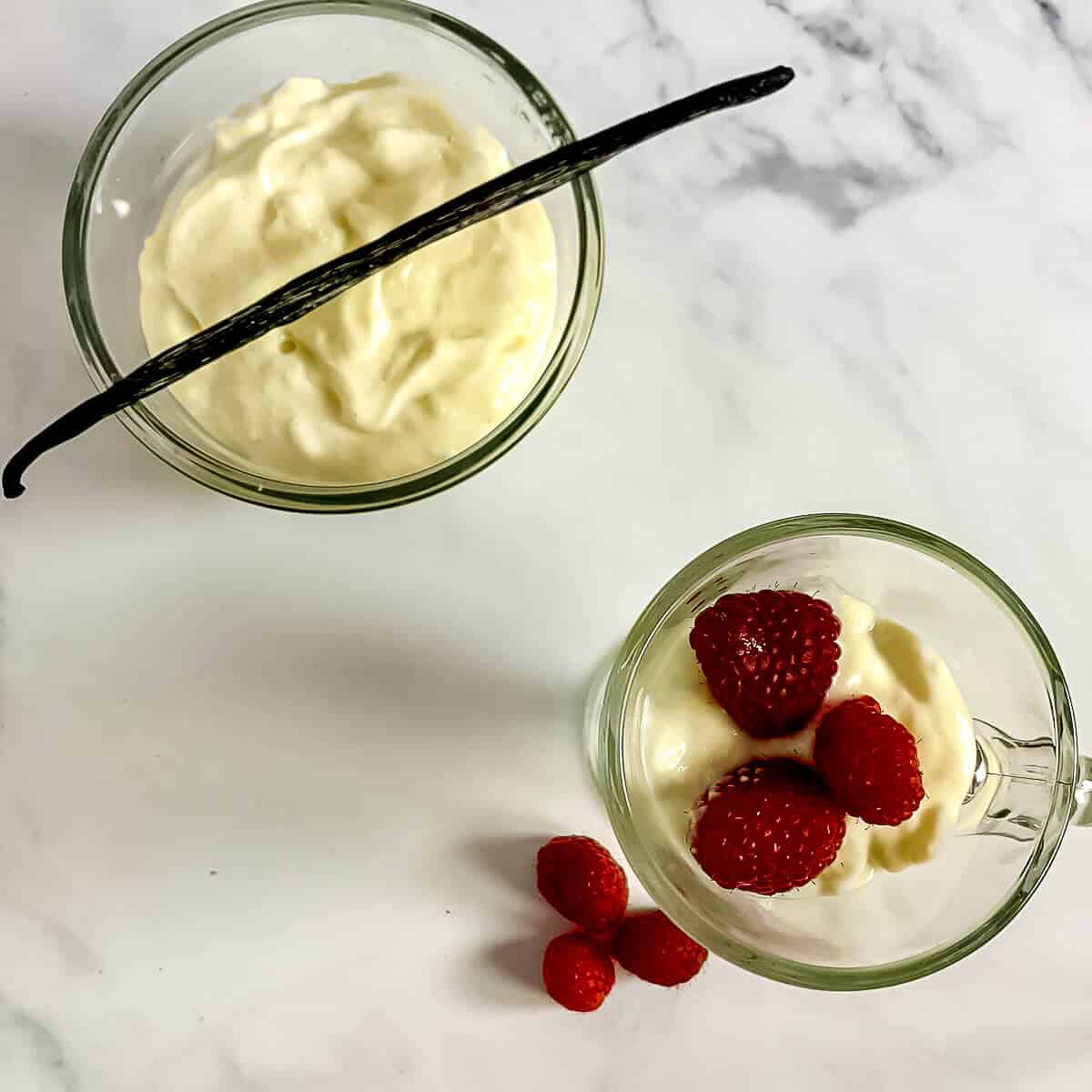 Vanilla pudding in a glass mug with raspberries next to another glass bowl of pudding with a vanilla bean from overhead
