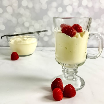 Vanilla pudding in a glass mug with raspberries in front of another glass bowl of pudding with a vanilla bean.