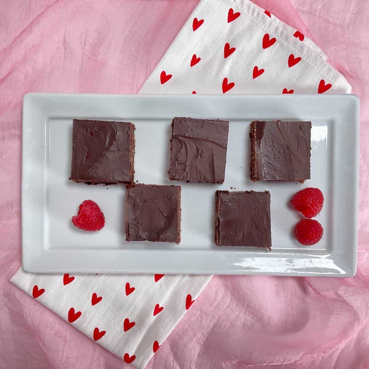 Five Chocolate Raspberry brownies & fresh raspberries atop a white plate on a white towel with red hearts on a pink scarf.