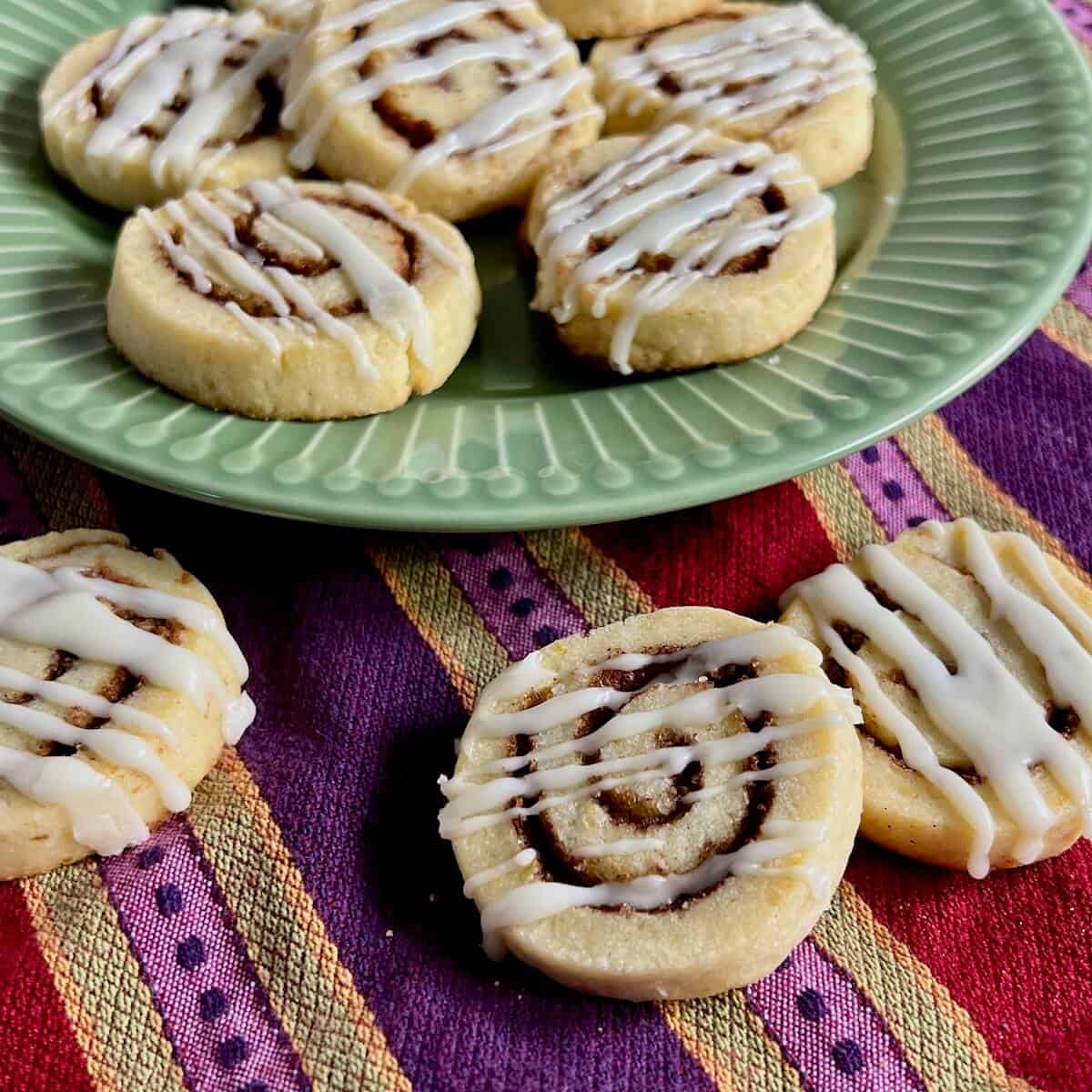 Three cinnamon roll cookies on a striped towel in front of more on a green plate.